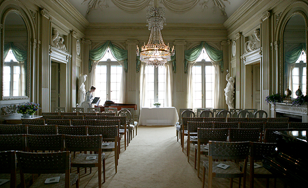 En tom sal på Gunnebo slott med tomma stolar och en kvinna i tidsenlig klädsel i ena hörnet.