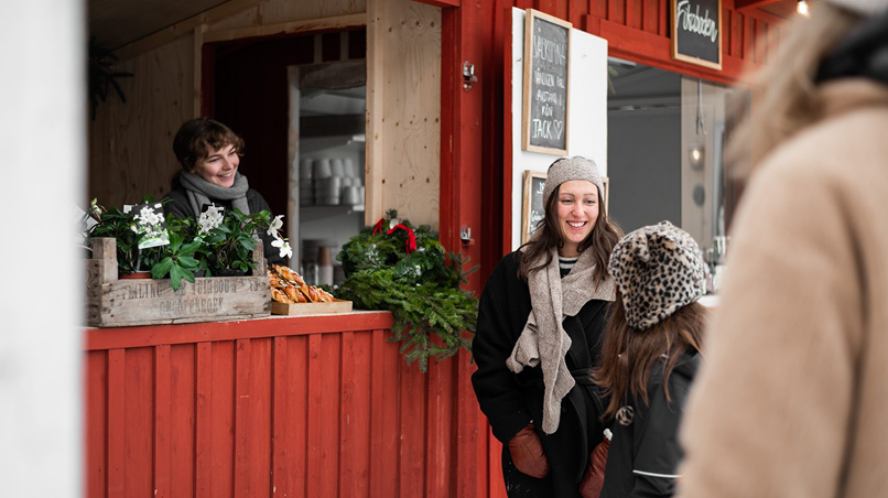 En kvinna står i ett marknadsstånd på Gunnebos julmarknad och ler mot besökarna som står framför henne.