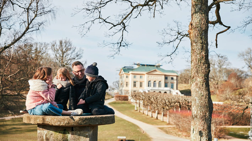 En man och tre barn sitter på en stenfigur med Gunnebo slott bakom sig.